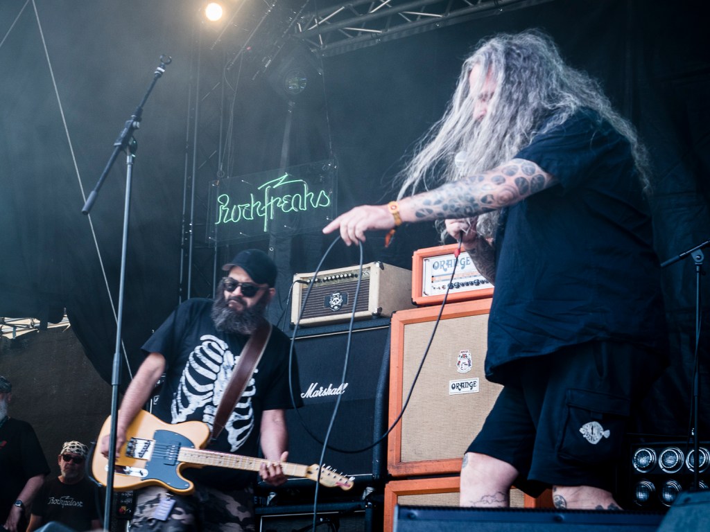 Zwei Musiker auf einer Festivalbühne, einer mit einer E-Gitarre und der andere mit einem Mikrofon, umgeben von Verstärkern und Bühnenbeleuchtung.