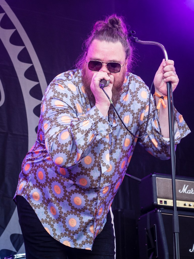 Ein Sänger auf der Bühne, der ein Mikrofon hält und eine modische Bluse mit buntem Muster trägt, während er mit Leidenschaft performt.