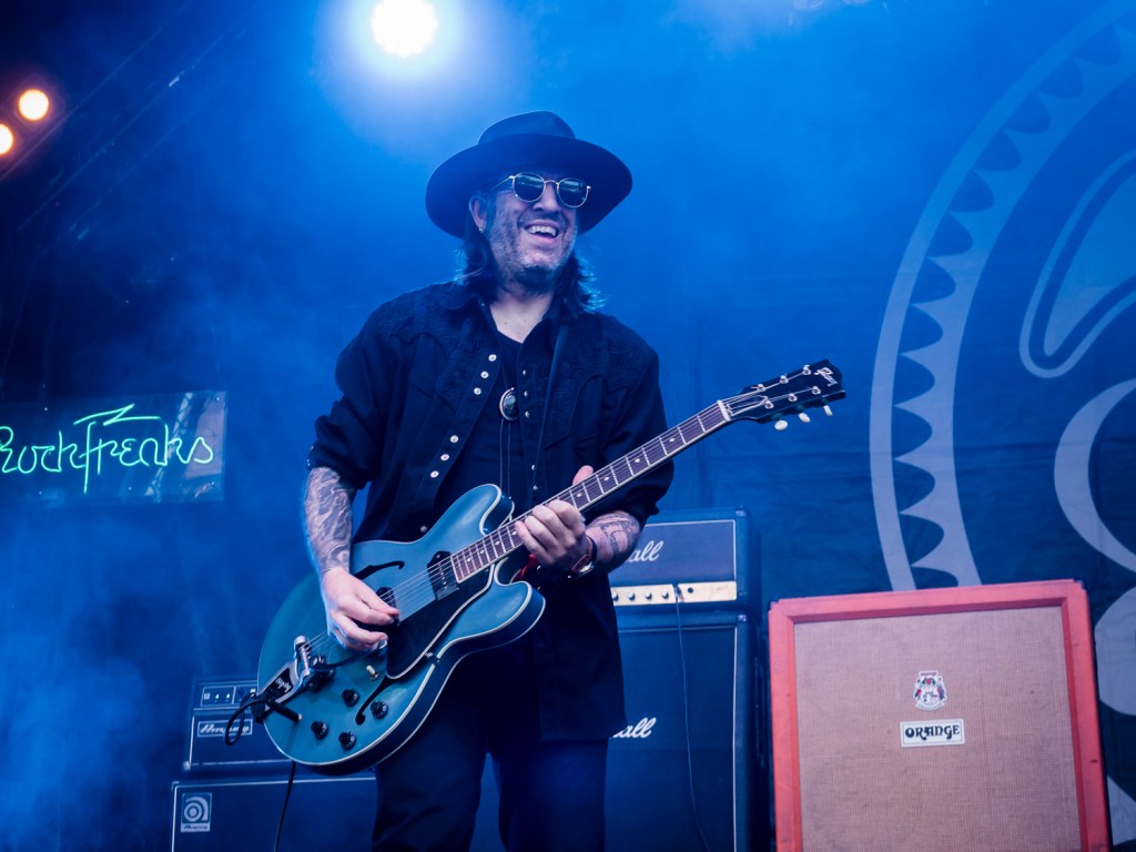 Ein Musiker auf der Bühne spielt E-Gitarre, während er lächelt und eine Sonnenbrille trägt. Im Hintergrund sind Verstärker und Bühnenbeleuchtung sichtbar.