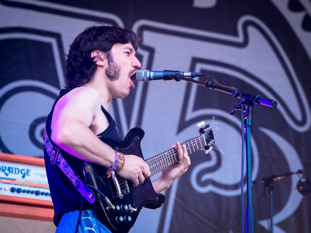 Ein Musiker singt leidenschaftlich ins Mikrofon, während er auf der Bühne eine E-Gitarre spielt.