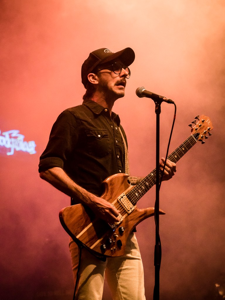 Ein Musiker steht auf der Bühne und spielt Gitarre, während orangefarbener Nebel im Hintergrund aufsteigt.