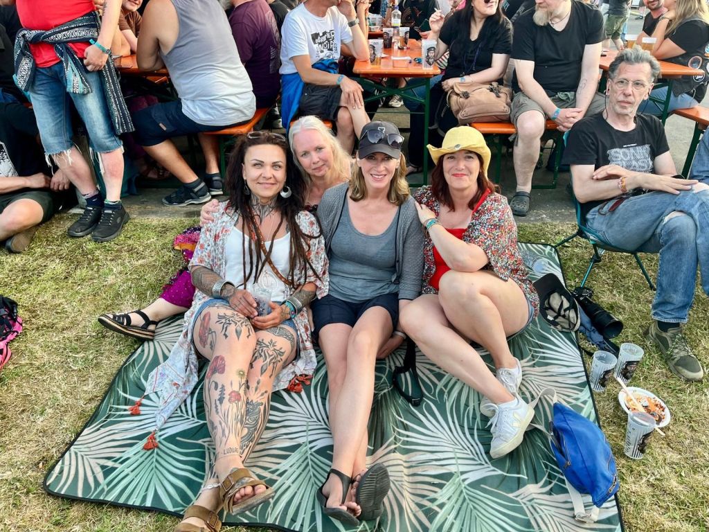 Gruppenshot von vier Frauen auf einer Picknickdecke, umgeben von Festivalbesuchern im Hintergrund, einige genießen Getränke. Die Frauen tragen lockere Kleidung und haben verschiedene Frisuren, während die Atmosphäre entspannt und fröhlich wirkt.