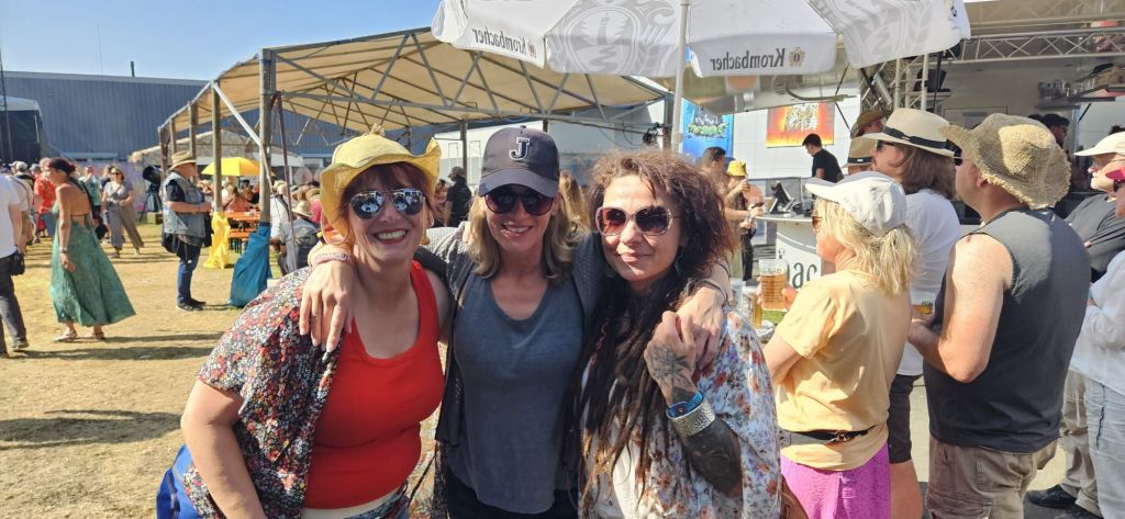 Drei Frauen, die lächeln und sich umarmen, stehen in einem Festivalgelände mit sonnigem Wetter und festlicher Atmosphäre. Im Hintergrund sind Schirme und Buden sichtbar, während andere Festivalbesucher in bunter Kleidung umherlaufen.