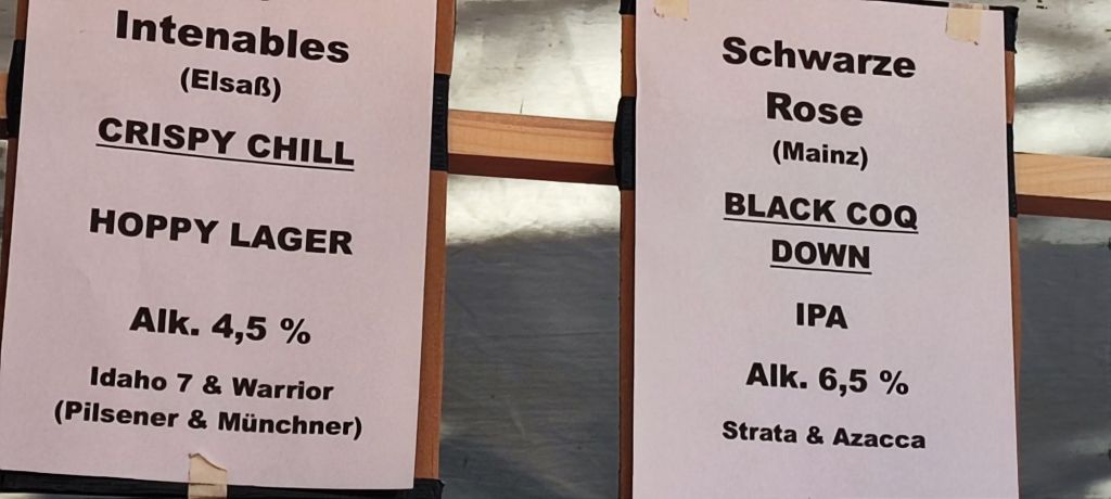 A sign displaying two craft beers: "Crispy Chill" Hoppy Lager from Intenables with 4.5% alcohol content, and "Black Coq Down" IPA from Schwarze Rose with 6.5% alcohol content, along with their ingredients.
